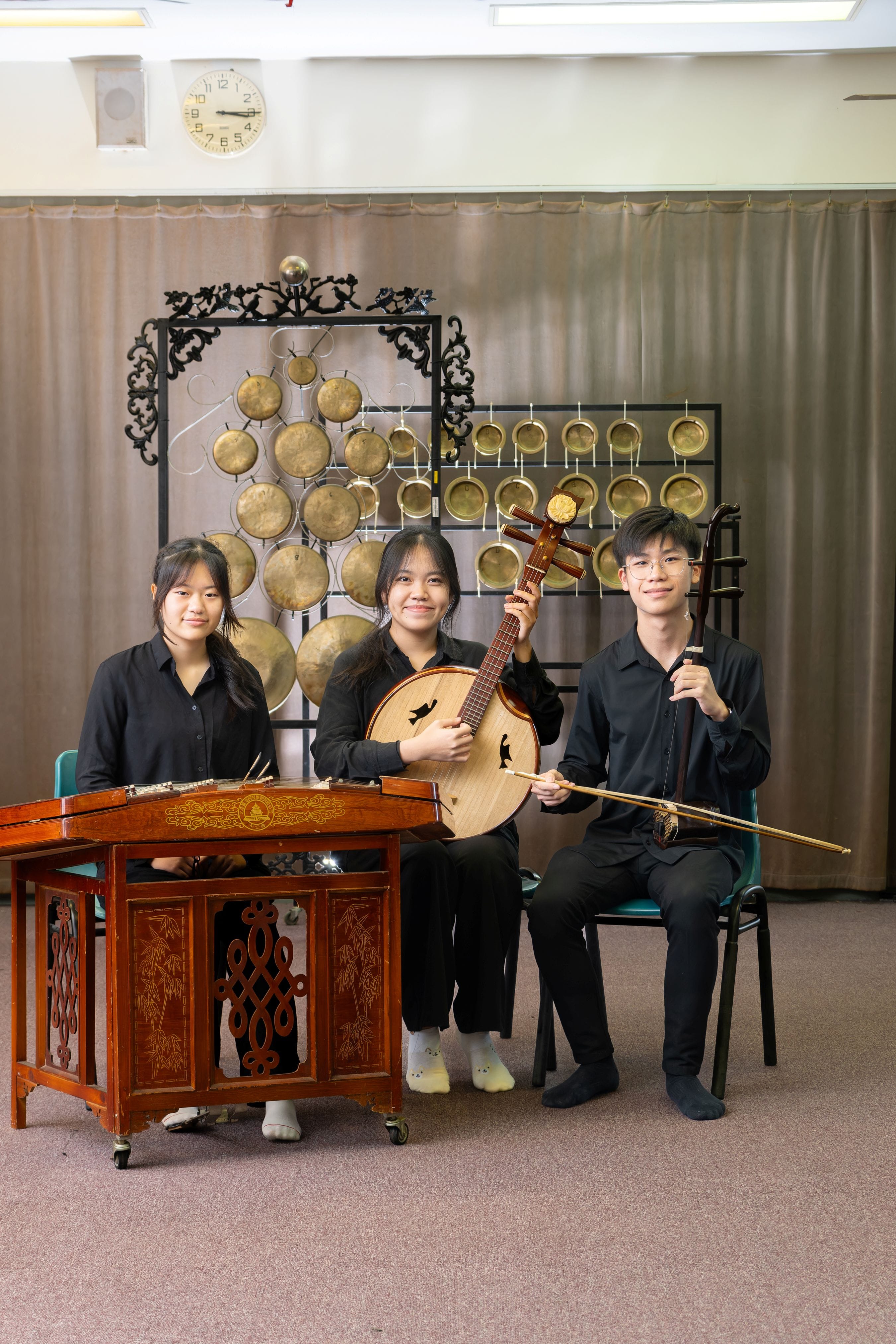 Three students in black attire pose indoors in front of a decorative rack of brass gongs. The student on the left sits behind a yangqin (a hammered dulcimer), the student in the centre holds a ruan (a round-bodied lute), and the student on the right holds an erhu (a two-stringed bowed instrument) and its bow.